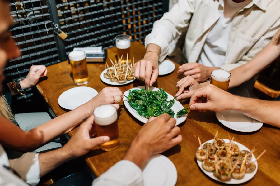 Friends share plates of tapas, including Padrón peppers, and glasses of beer at a wooden table in a bar.
