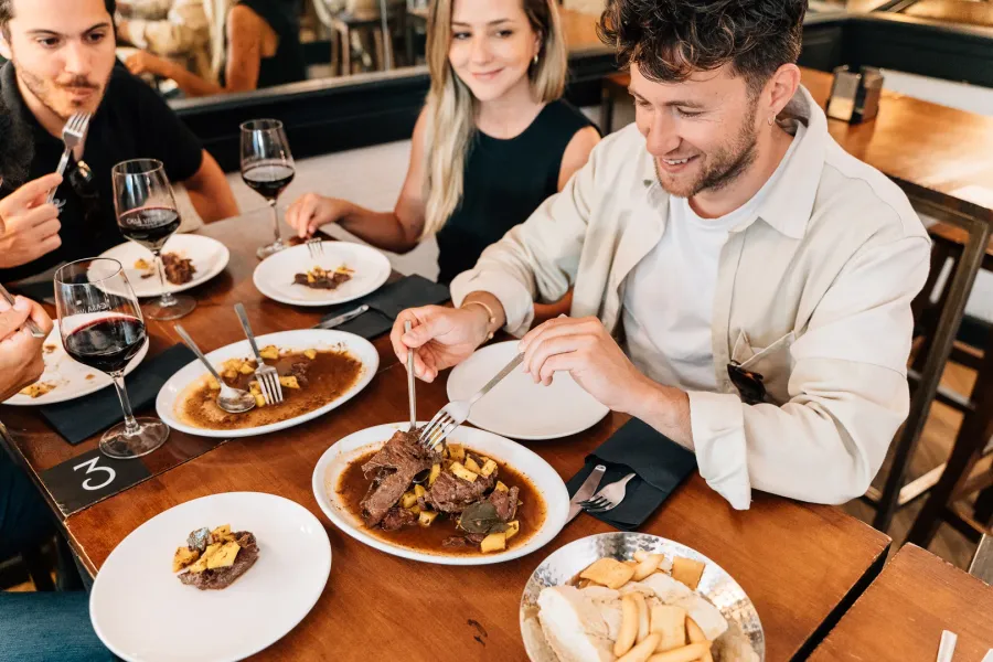 Three people at a wooden table sharing plates of beef stew and drinking glasses of red wine in a restaurant.