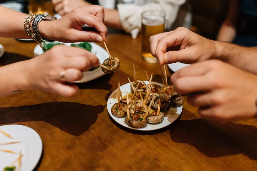 Hands picking up mushroom tapas with toothpicks from a plate on a wooden table.