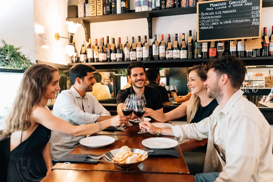 A group of five friends sits at a table in a bar, smiling and toasting with glasses of red wine.