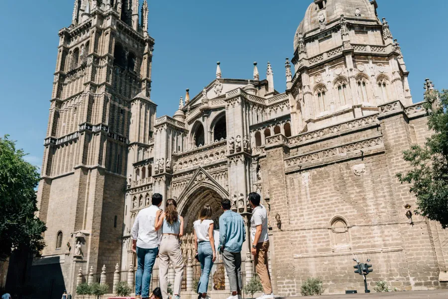 A group of five people, seen from behind, look up at the intricate stone facade of the Toledo Cathedral on a sunny day.