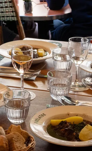 A dining table set with plates of stew, one reading 'ADÉLAÏDE', glasses of water and rosé wine, and a bread basket.