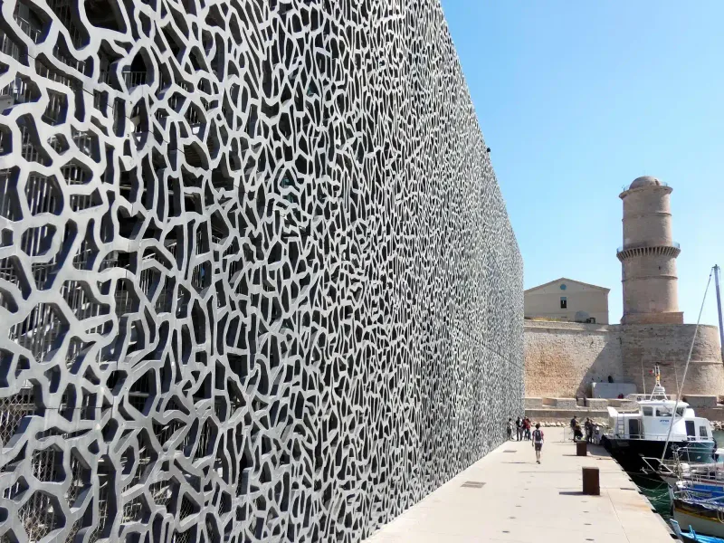 Modern building with intricate, net-like facade and old stone fort under a clear blue sky on a sunny day in Marseille.