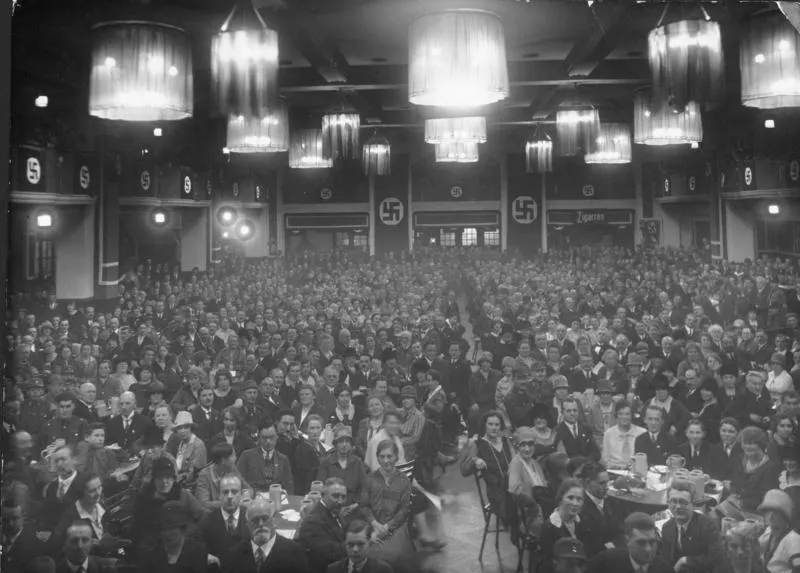 Black and white photo of a large crowded hall filled with people seated at tables. Swastika banners and 'Zigaretten' sign ...