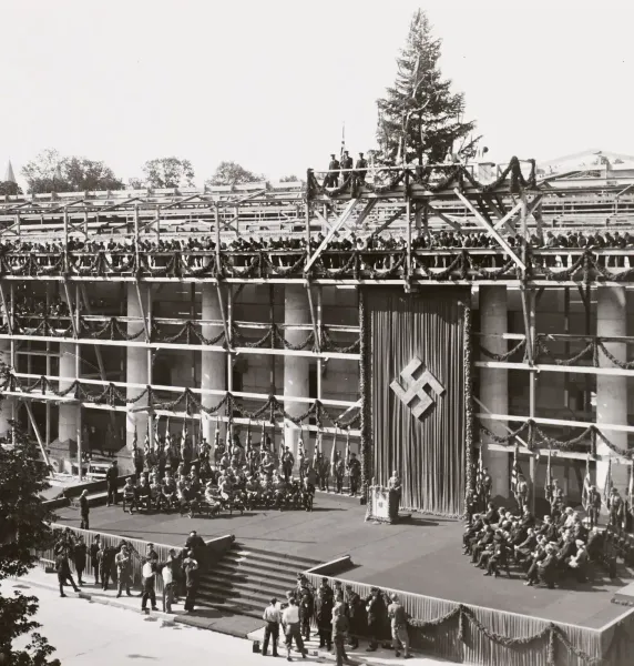 Black and white photo of a building under construction, decorated with garlands and a large swastika banner. People are ga...