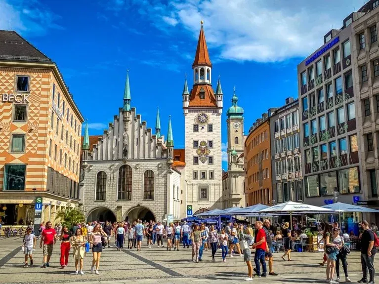 Crowded Marienplatz in Munich with the Old Town Hall and the tower of the New Town Hall under a blue sky.