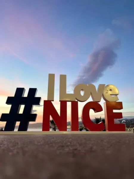 Large '#ILoveNICE' sign illuminated at dusk, with a soft pink and blue sky over the city and sea in Nice.