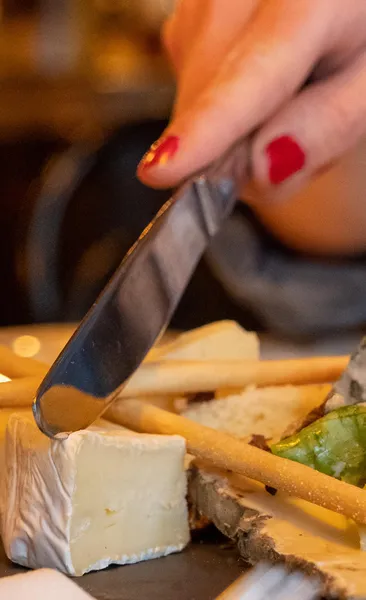 Close-up of a hand with red nail polish cutting a slice of white-rinded cheese on a plate with breadsticks.