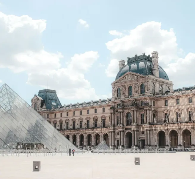 The Louvre Museum palace building with a dark dome and the glass pyramid in the foreground under a cloudy sky.