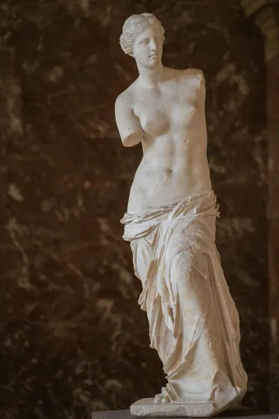 White marble statue of Venus de Milo with no arms, partially draped, standing against a dark brown marbled wall.