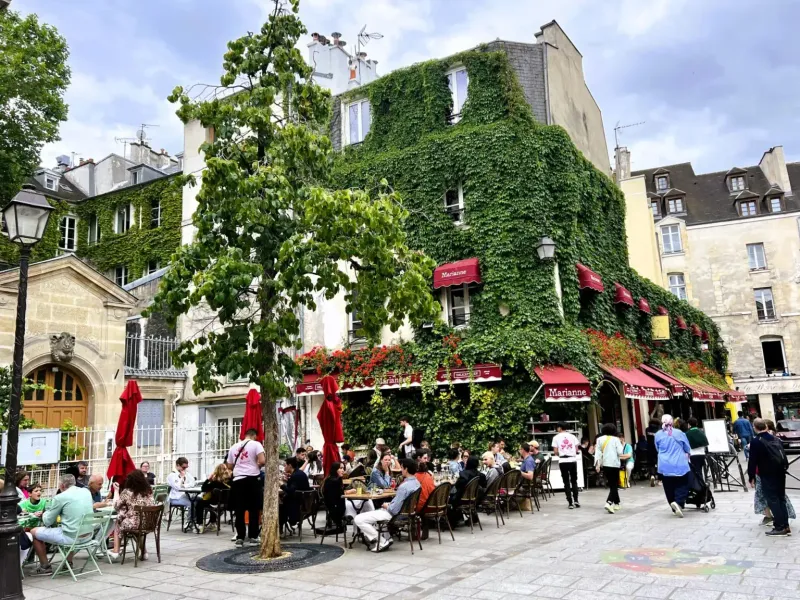 Building covered in green ivy and red awnings, with an outdoor restaurant patio in Paris. People are dining under a cloudy...