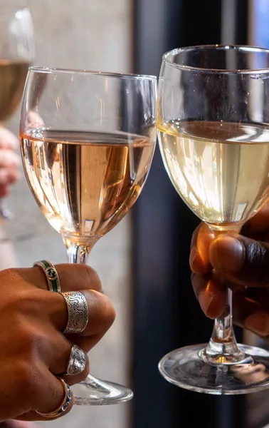 Two hands holding wine glasses, one with rosé wine and the other with white wine, clinking in a toast. One hand wears rings.