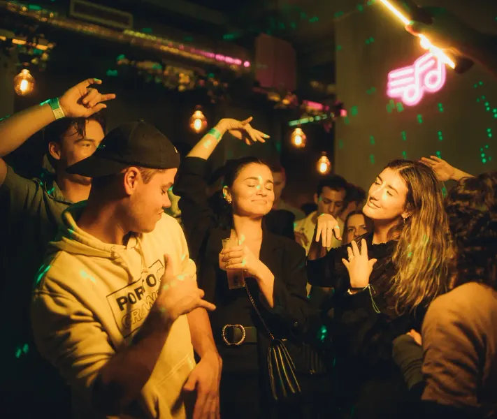 People dancing in a dimly lit club or bar, with green laser lights and a pink neon musical note sign.