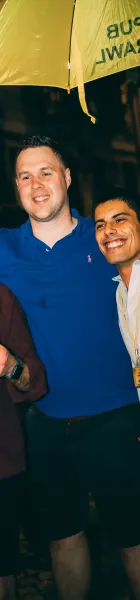 Two smiling men under a yellow umbrella with inverted 'PUB CRAWL' text, in a nighttime setting.