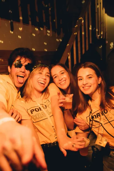 Four young people, two men and two women, smiling in a dimly lit bar. They wear yellow t-shirts with 'PORTO PUB CRAWL' and...