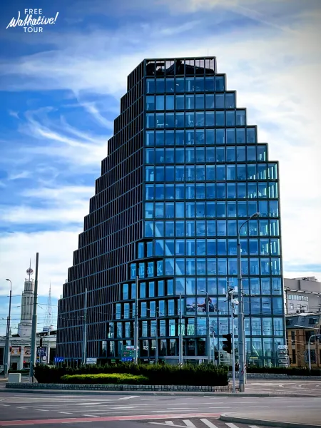 Modern Bałtyk building in Poznań with a stepped glass facade, set against a blue sky, with a street and historical tower v...