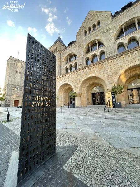 Dark monument with gold numbers and 'HENRYK ZYGALSKI' on a paved plaza, facing Poznan Imperial Castle with arches.