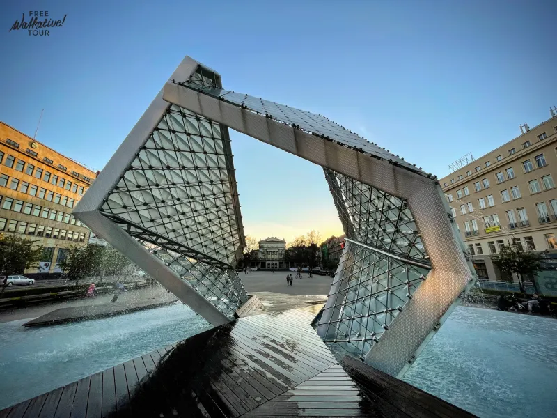 Modern glass and metal fountain with water jets, shaped like an open cube, in a city square at dusk. Buildings in the back...