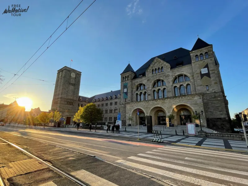 Stone Imperial Castle building in Poznań with a clock tower, tram tracks, and a crosswalk in the foreground. Sun is low on...
