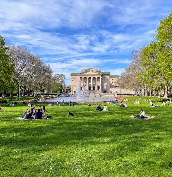 Grand Theatre building, fountain, and people relaxing on green grass in Poznań park on a sunny day.
