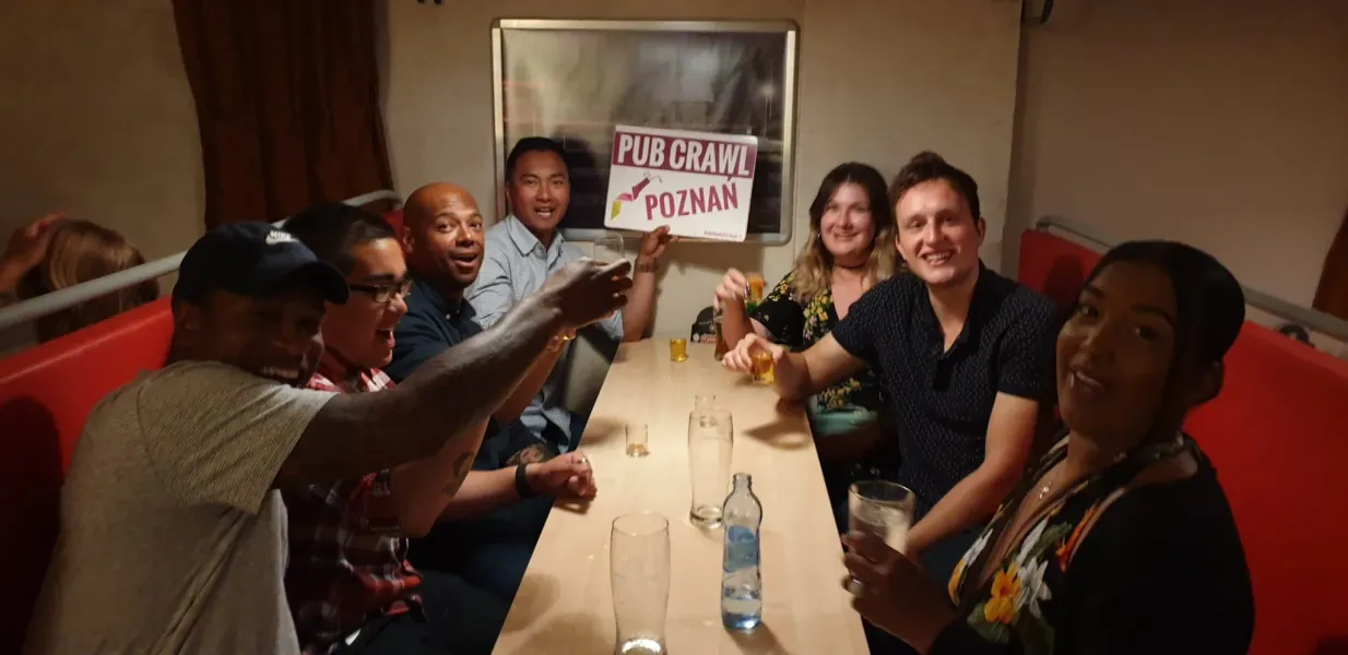 Diverse group of young adults smiling at a table in a pub, some holding drinks. One person displays a 'Pub Crawl Poznan' s...