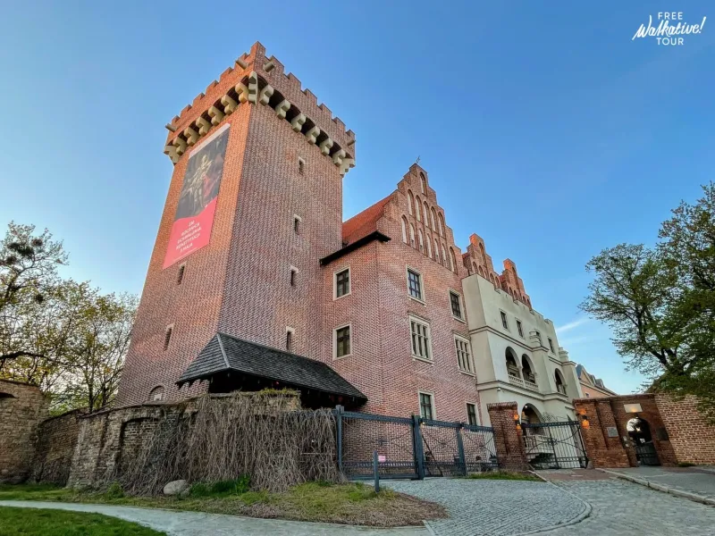 View of the Royal Castle in Poznań, a large brick building with a tall tower, gabled facade, and old wall, under a blue sky.
