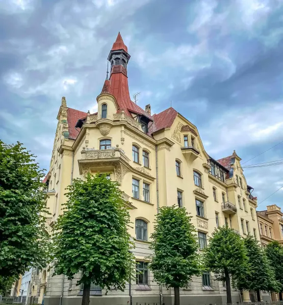 Ornate Art Nouveau building in Riga with a yellow facade, red roof, and central spire, framed by green trees against a clo...