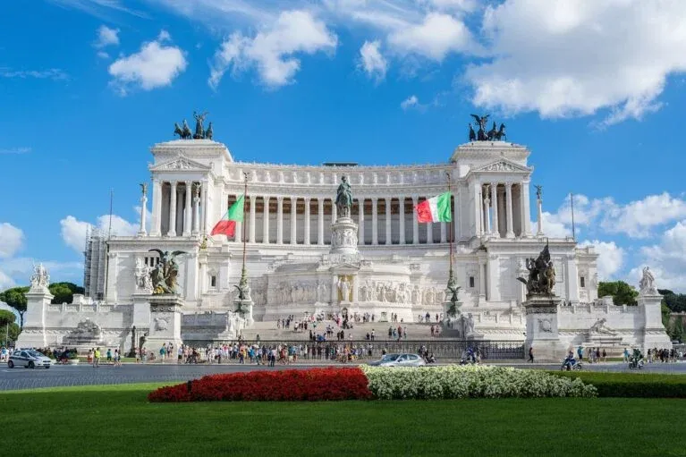 Grand white marble Altar of the Fatherland in Rome under a blue sky with clouds. Italian flags fly on either side. People ...