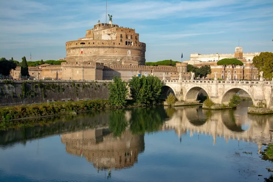 Castel Sant'Angelo and Ponte Sant'Angelo spanning the Tiber River under a blue sky, with reflections on the water.