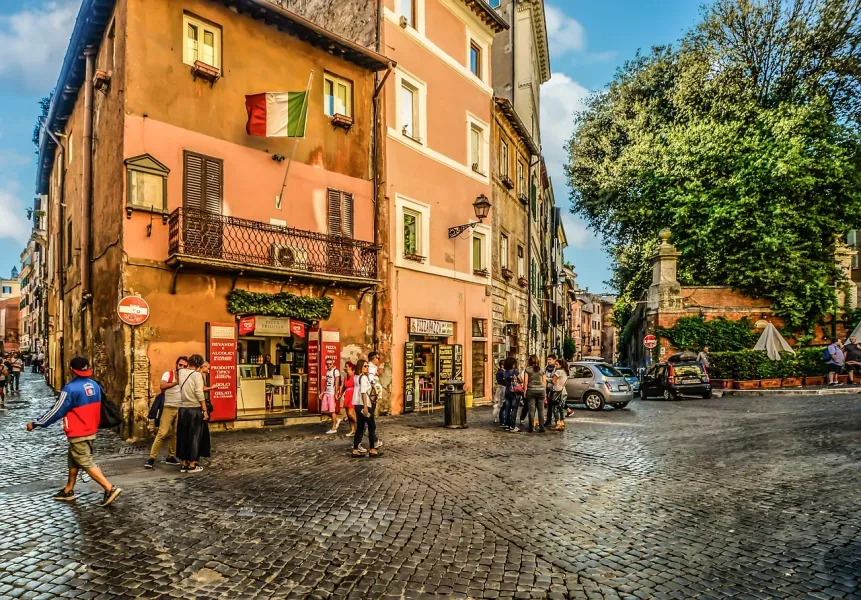 Cobblestone street in Rome with historic buildings, an Italian flag, people walking, and parked cars under a blue sky.