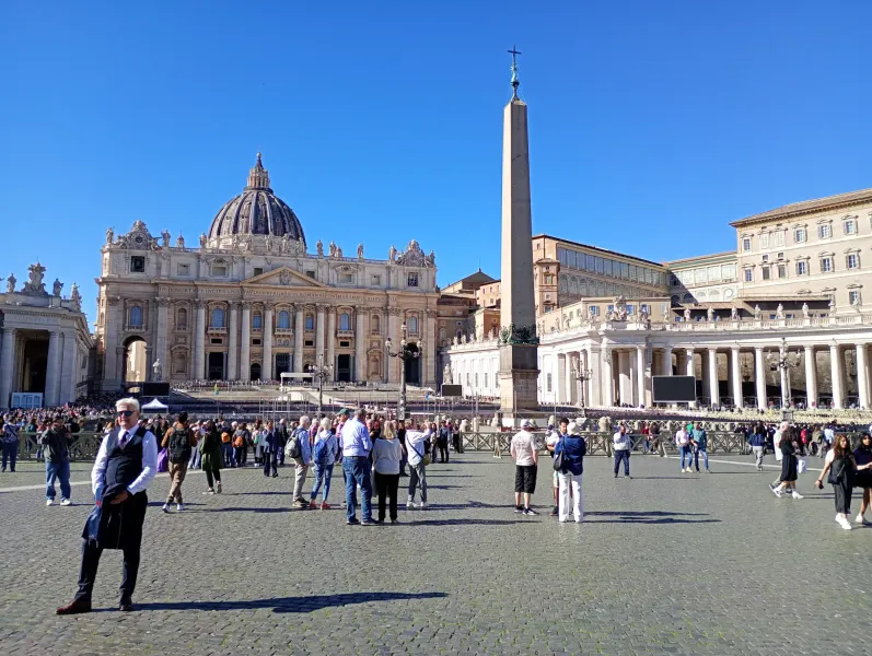Tourists walking in St. Peter's Square with the central obelisk and St. Peter's Basilica facade behind.