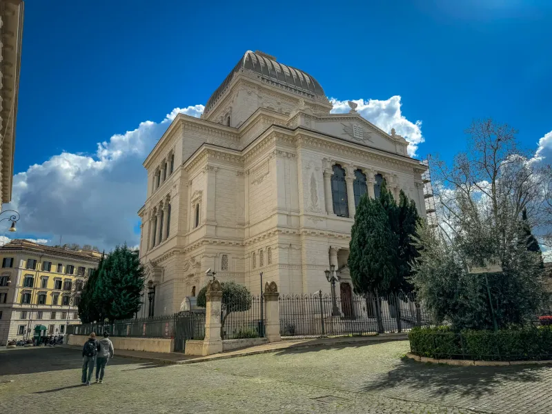 The Great Synagogue of Rome with its dome, a cobblestone street, green trees, and two people walking under a bright blue sky.
