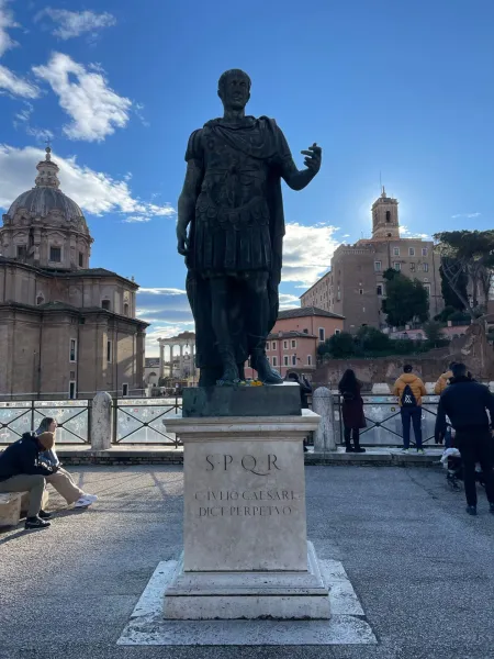 Bronze statue of Julius Caesar in Rome, with Roman Forum ruins and a domed church under a blue sky.