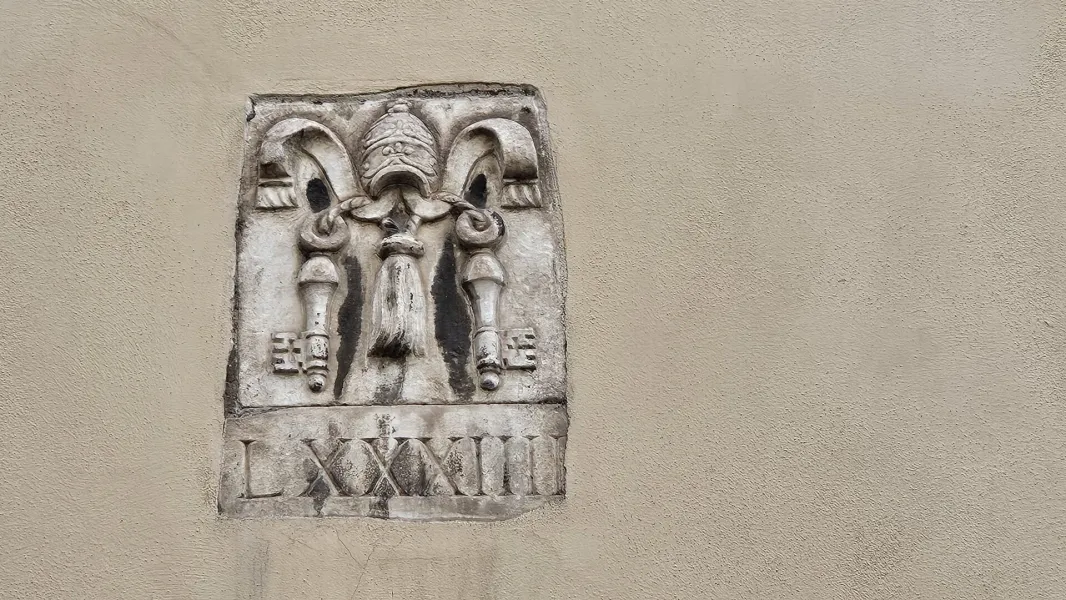 Stone relief on a textured wall showing a papal tiara, crossed keys, and tassels, with Roman numerals LXXXIII below.