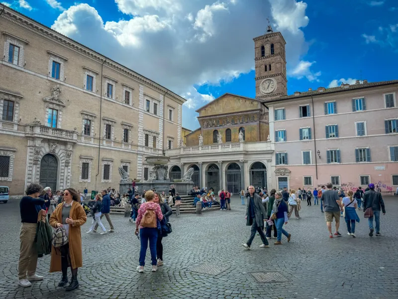Cobblestone Piazza Santa Maria in Trastevere, Rome, with a basilica, bell tower, fountain, and many people walking.