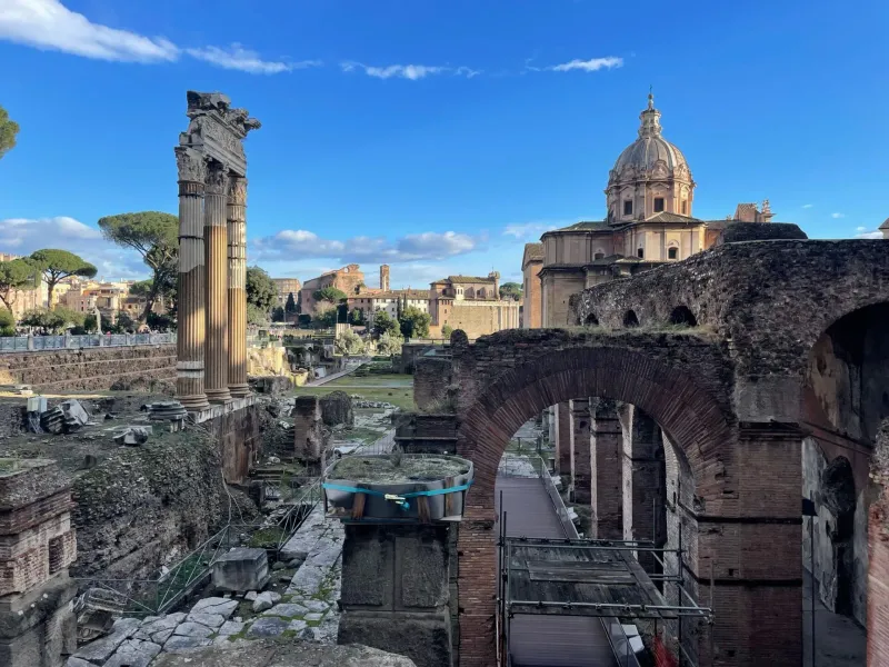 Ancient Roman Forum ruins with the Temple of Saturn columns, other temples, and scattered visitors under a blue sky.