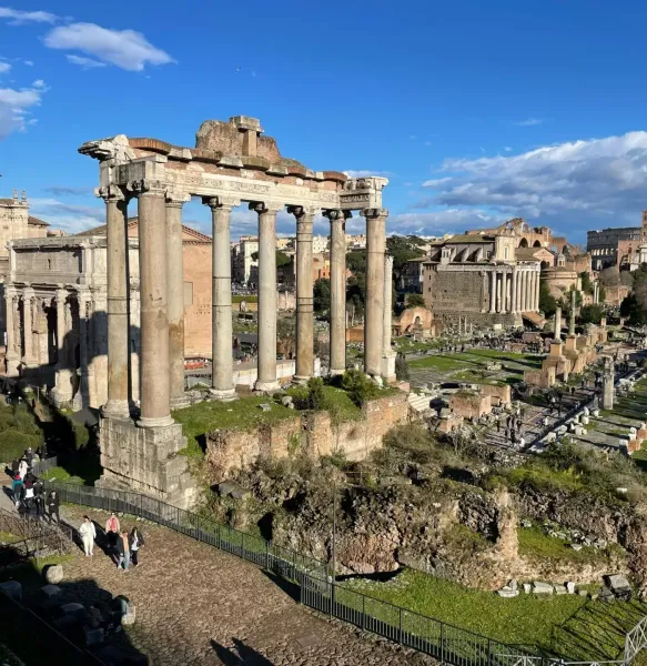 View of the Roman Forum in Rome, showing ancient ruins including eight tall columns of the Temple of Saturn under a blue sky.