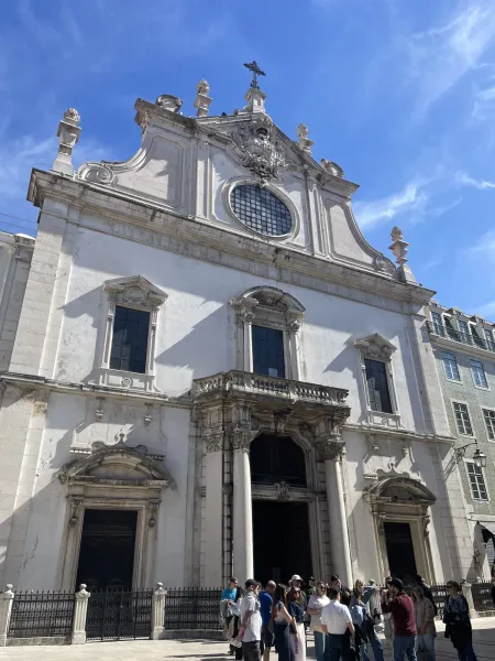 Ornate white facade of São Domingos Church in Lisbon, with large dark doors, a balcony, and a cross on the roof. People ga...