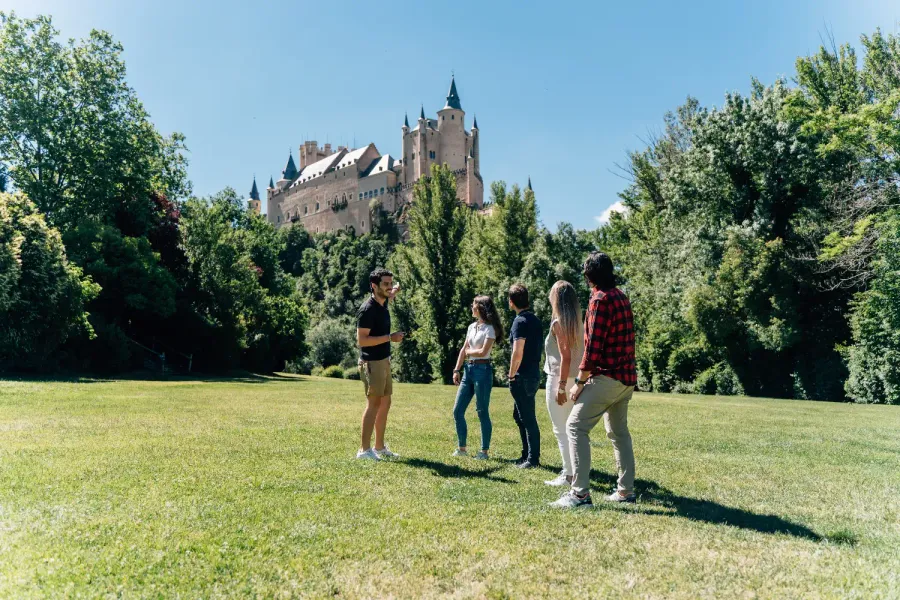 A tour guide speaks to a small group in a park with the Alcázar of Segovia castle in the background.