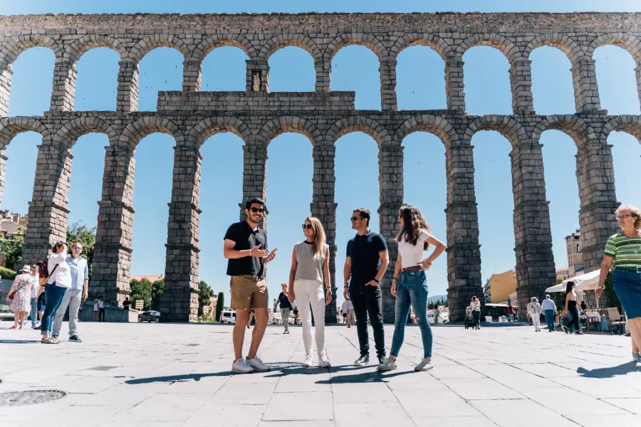 A tour guide talks to three tourists in front of the large, sunny Aqueduct of Segovia in Spain.
