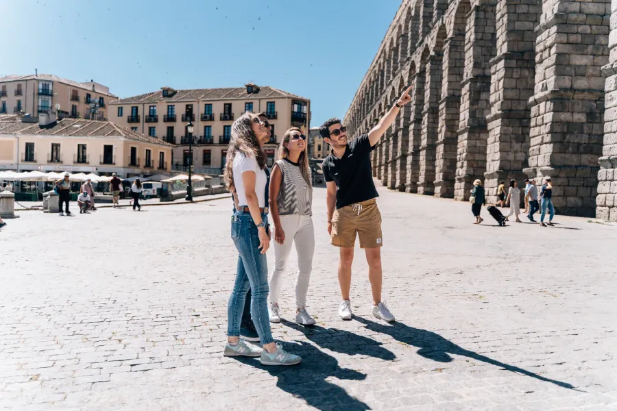 A tour guide points up at the Aqueduct of Segovia for three tourists on a sunny day.