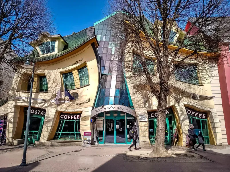 The Crooked House (Krzywy Domek) in Sopot, with a wavy facade and green roof, shops on the ground floor, and bare trees.