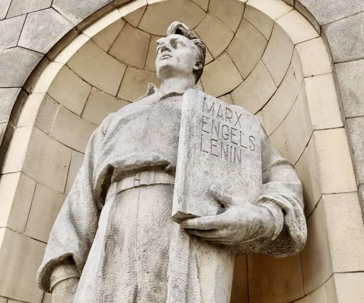 Close-up of a weathered stone statue holding a book inscribed with 'Marx Engels Lenin'.