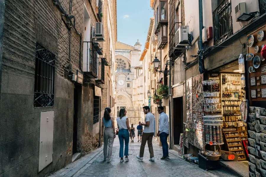 Four young people walk down a narrow alley in Toledo, with the cathedral in the background and a souvenir shop on the right.