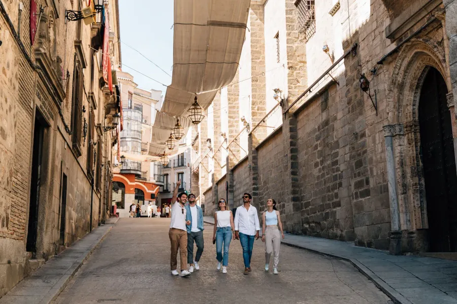 A group of five young people walks down a narrow stone street in Toledo, shaded by traditional awnings.