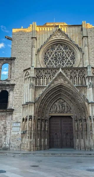 Gothic stone facade of Valencia Cathedral with a large rose window, ornate arched portal, and sculpted figures under blue ...