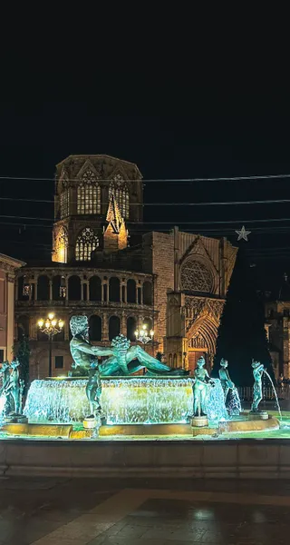 Illuminated Turia Fountain with bronze statues and the illuminated Valencia Cathedral at night. A large Christmas tree is ...