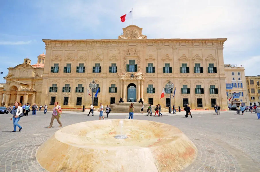 The Auberge de Castille, an ornate Baroque building with green shutters and the Maltese flag, stands on Castille Square wi...