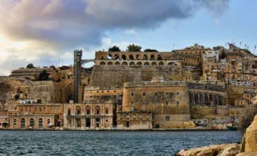 View of Valletta's historic fortifications and stone buildings along the Grand Harbour, with a dramatic cloudy sky above.