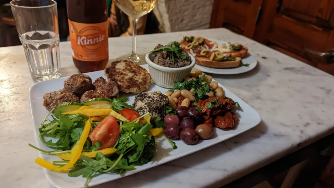 A platter of Maltese food including meatballs, salad, beans, olives, and cheese, with a Kinnie bottle and wine glass on a ...
